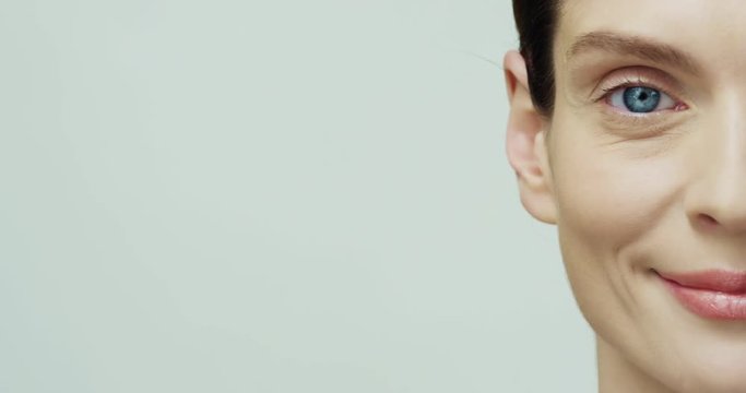Close up of the half face of the young charming happy Caucasian lady with blue eyes and smooth skin looking straight to the camera and smiling on the white background.