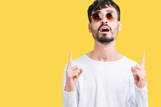 Young handsome man wearing sunglasses over isolated background amazed and surprised looking up and pointing with fingers and raised arms.