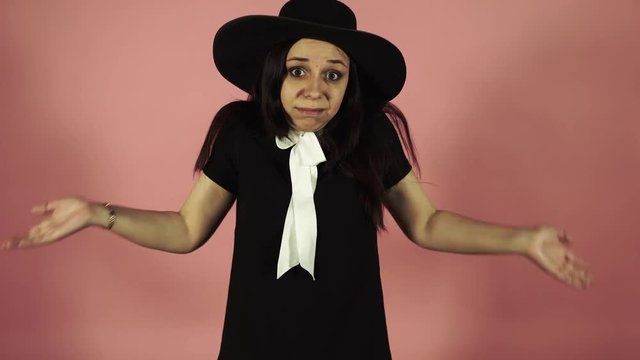 Young female in black dress and hat on a pink background. Attractive woman wearing black outfit standing on pink background in perplexity on a pink background. The confused woman shrugs