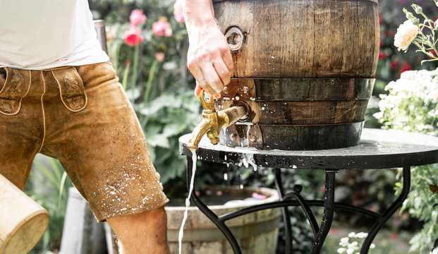 bayerischer Mann in Lederhose sticht ein Holzfass Bier im Garten an und genießt den ersten Schluck