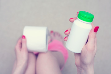 Female legs, girl sitting on the toilet, holding in hand white container of medicine, toilet paper,...