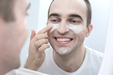 Attractive man, looking in the mirror in the bathroom, cosmetic mask, smiling, close up