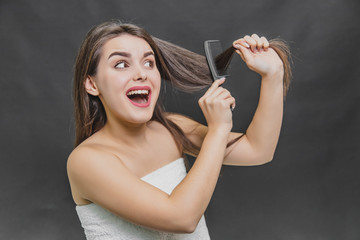 Young pretty girl brushing her hair and looking for hair falling on the comb and hands on a black background. Hair care, pain when combing. A frightened girl lifting her hair, opening her mouth.