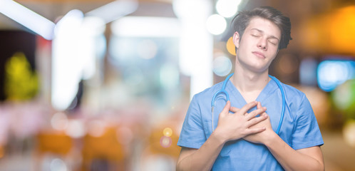 Young doctor wearing medical uniform over isolated background smiling with hands on chest with closed eyes and grateful gesture on face. Health concept.