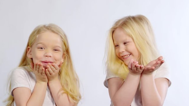 Chest-up shot of adorable blonde twin girls with long hair blowing kisses at camera, smiling and laughing, while posing in studio on white background