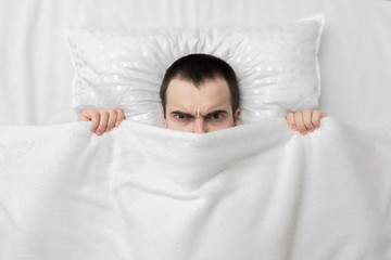 Gloomy guy in the bedroom, hiding under white blanket, man is angry and does not want to get up so early in the morning, top view