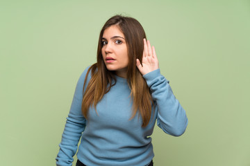 Young girl over green wall listening to something by putting hand on the ear