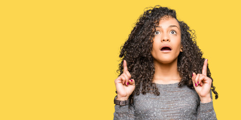 Young beautiful woman with curly hair wearing grey sweater amazed and surprised looking up and pointing with fingers and raised arms.