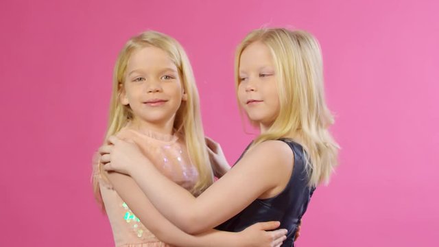 Waist-up shot of blonde 6-year-old Caucasian twin girls with long hair, wearing sparkly party dresses, hugging and looking at each other, then turning to camera and smiling, posing on pink background