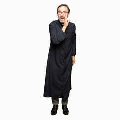 Middle age priest man wearing catholic robe hand on mouth telling secret rumor, whispering...