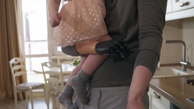 Tilting medium shot of multitasking amputee father holding up young daughter with myoelectric arm and preparing vegetable salad for lunch while talking on phone pressed between ear and shoulder