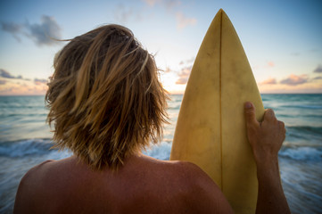 Sunset silhouette of a surfer with blond hair holding his surfboard in golden sun on the shore of the beach