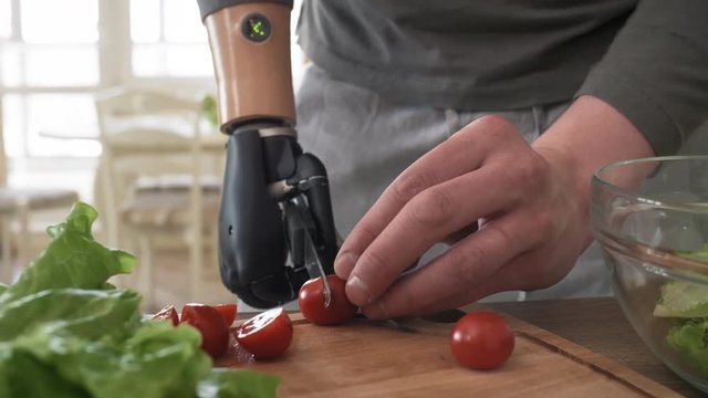 Close-up hands shot of self-sufficient male amputee gripping knife with bionic prosthetic forearm and chopping cherry tomatoes with precision and dexterity to make vegetable salad