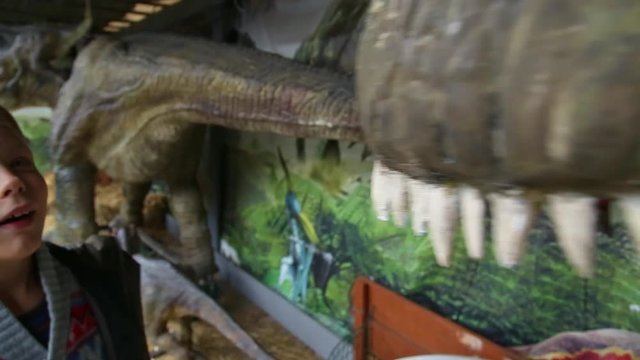 Young happy kid looks at teeth of model of huge scary dinosaur at theme park outdoor. Real time full hd video footage.