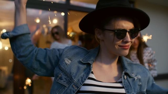 Medium close-up of 20-something hipster brunette wearing black fedora, denim jacket and sunglasses, dancing at house party holding a sparkler, with her girlfriends in background