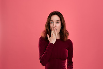 Surprised young lady standing isolated over dark pink background with hands raised close to mouth expressing emotion of shock.