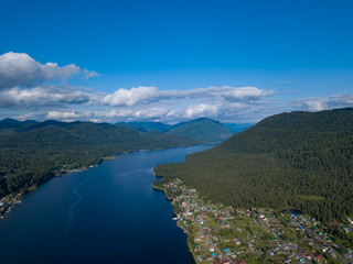 Aerial view of mouth at wide Teletskoye lake in the Altai Mountains by the blue water, sky with white clouds, green trees on the slopes of the rocks and village on shore. Rest and travel in nature.