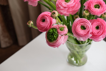 Pink Ranunculus bouquet in transparent glass vase on white table. Close-up. For flower delivery, social media. Soft selective focus. Copy space. Horizontal