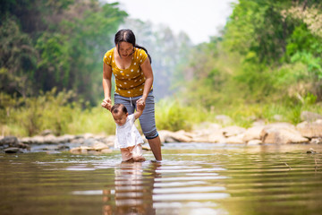 Asian woman holding her baby girl's hands to practice walking in water with beautiful forest view in background