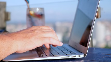 male typing on a laptop in a cafe on the roof of a high-rise with a beautiful panoramic view of the city, close up