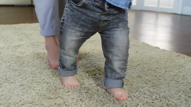 Close-up feet shot of toddler taking his faltering first steps while being guided by his mother, walking barefoot across shaggy beige carpet, dressed in turned-up jeans