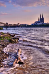 Köln Stadtportrait bei Sonnenuntergang, starken Wellen im Vordergrund, Kölner Dom und Hohenzollernbrücke im Hintergrund. Kölner Skyline Poster und Postkartenmotiv im Hochformat. © 77pixels