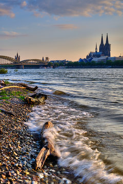 K&ouml;ln Stadtportrait bei Sonnenuntergang, starken Wellen im Vordergrund, K&ouml;lner Dom und Hohenzollernbr&uuml;cke im Hintergrund. K&ouml;lner Skyline Poster und Postkartenmotiv im Hochformat.
