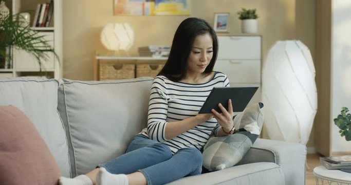 Cheerful Asian Young Woman In The Striped Shirt Scrolling And Tapping On The Tablet Device On The Couch In The Living Room.