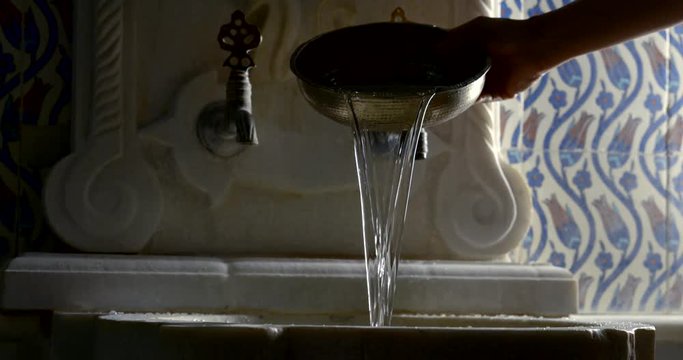 human is pouring water from bronze cup into stone washbasin in dark room, close-up