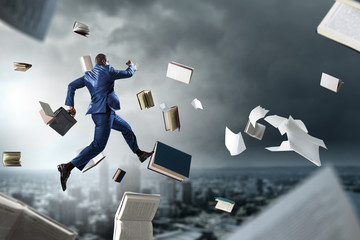 Back view of black businessman with briefcase running among flying books and papers on dark cloudy...