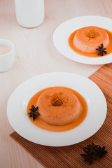 Pudim de leite com calda de caramelo com uma flor de anis estrelado seca ao lado em prato branco raso, sobre uma esteira de bamboo