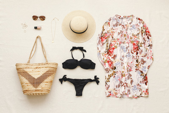 Black bikini swimsuit, floral shirt dress, straw boater hat, wicker beach bag, sunglasses, gold necklace, rings on beige background. Woman's swimwear and beach accessories. Flat lay, top view, outfit.