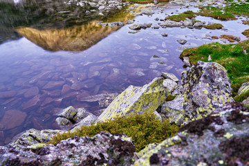 Rocks with green lichens, purple sunset light, and a mountain peak reflecting in Lake Bucura, Retezat Mountains (part of Carpathians), Romania. Summer tourism concept in Europe.
