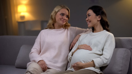 Young woman sitting on sofa with her pregnant friend, happy future mother