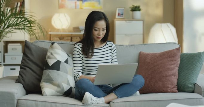 Asian pretty young woman freelancer working over her project on the laptop computer and thinking while sitting on the couch at home.