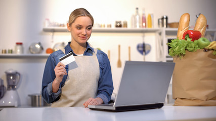 Girl in kitchen holding bank card, satisfied with food purchase over internet