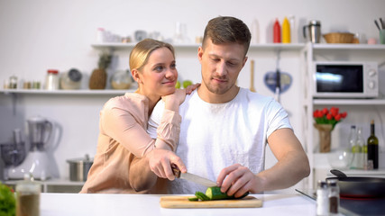 Husband slicing vegetable, loving wife embracing him, romantic moment in kitchen