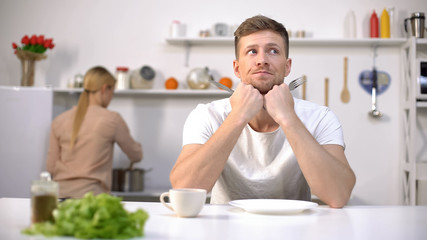Anxious husband waiting for lunch, wife cooking on background, lack of time