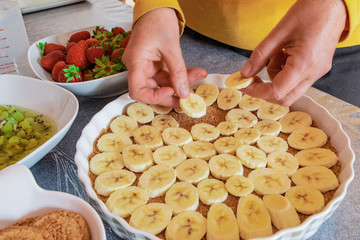 cooking fruit cake. Female hands spread bananas in a baking dish. On the background are strawberries, kiwi and cookies.