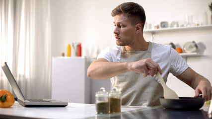 Bachelor preparing meal in pan, reading recipe and cooking tips online, courses