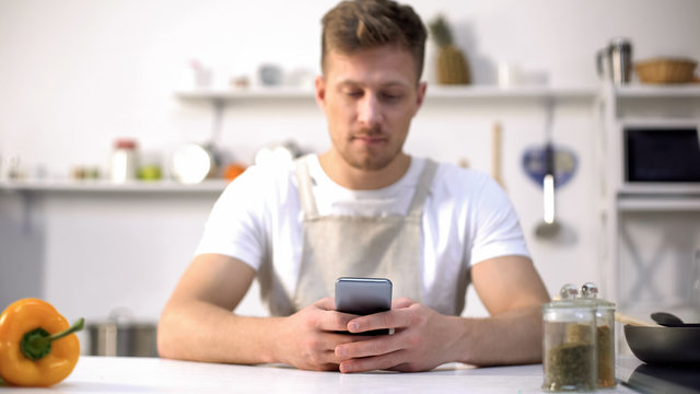 Young man in apron searching food recipe in cellphone, tips in culinary blogs