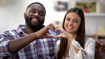 Joyful mixed-race couple showing heart sign made with hands at camera, love