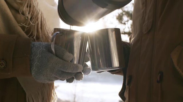 Close up hands shot with lens flare of couple enjoying hot drinks in snowy winter forest against bright setting sun