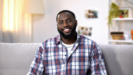 Obraz premium Cheerful African-American man sitting on sofa and looking at camera, relaxing