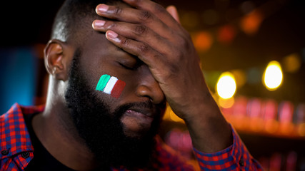 Anxious black soccer fan with italian flag painted on cheek making facepalm loss
