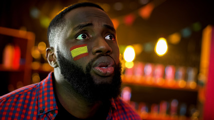 Anxious african american man with spanish flag on cheek supporting sport team