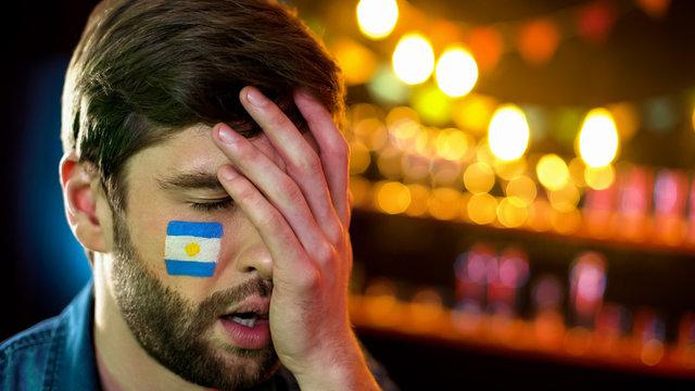 Argentinian football fan with flag on cheek making facepalm, unhappy with loss