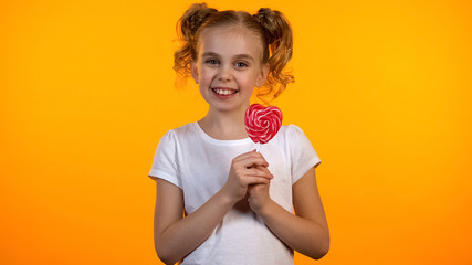 Pretty candy girl holding heart-shaped lollipop and smiling to camera, happiness