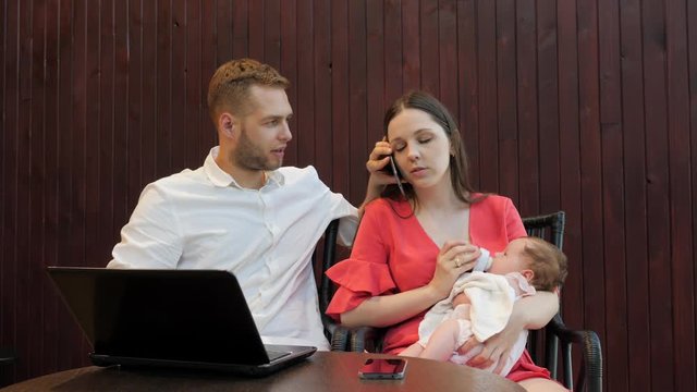 Busy parents feeding their baby infant sucking milk from bottle with nipple while adults have phone talk and using laptop computer online shopping