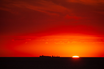 Cargo ship on the sea in sunset with cloud formations in red light. Sun touches the sea and is almost gone.
